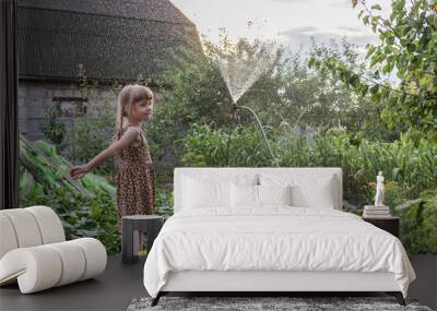 A little girl with pigtails in a sundress stands under the spray of a watering pad in the middle of a vegetable garden in summer Wall mural