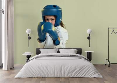 Portrait of little karate girl in white kimono wearing gloves and protective helmet, looking focused at camera, ready to punch, doing martial arts isolated over green background Wall mural