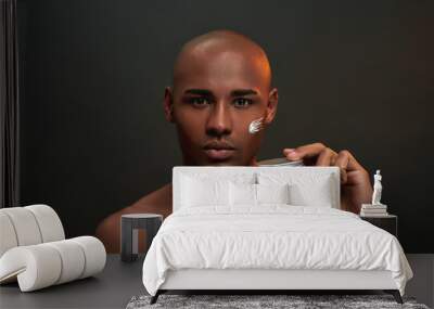 Portrait of shirtless african american man looking at camera, holding beauty product while posing with cream applied on his cheek isolated over black background Wall mural