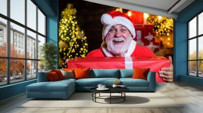 Present box. New Year eve. Santa Claus holds present. Smiling man in Santa hat hold present gift box. Santa man with Christmas gift box. Merry Christmas and happy New year. Wish you merry Christmas. Wall mural