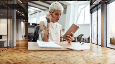 Skype call. Portrait of young cute blonde tattooed business woman putting on wireless earphones and using her touchpad while sitting in the modern office Wall mural