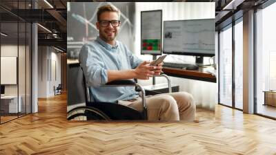 Successful trader. Portrait of young and cheerful male businessman in a wheelchair holding his smartphone and smiling while sitting at his workplace in modern office. Disabled people Wall mural