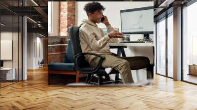 Successful young male trader talking on the phone, discussing stock market while sitting at desk in the living room at home Wall mural