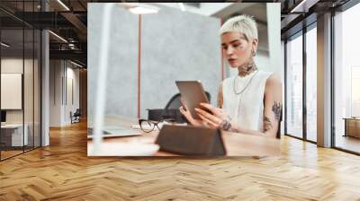 Surfing the net. Portrait of young stylish blonde tattooed business woman using touchpad while sitting at her working place in the modern office. Digital technologies at work Wall mural