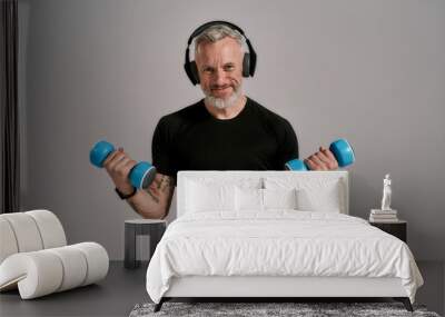 Train more. Middle aged muscular man in black t shirt and headphones smiling at camera holding blue dumbbells, posing in studio over grey background Wall mural