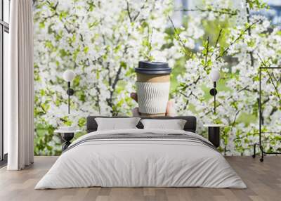 A woman's hand holds a disposable cup of coffee against the blossoming spring tree. The beginning of a beautiful spring day. Good mood and lifestyle Wall mural