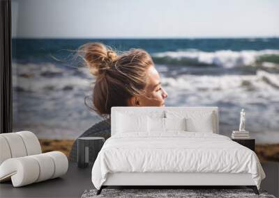 A young beautiful Caucasian girl with a bun of hair on her head in a white boho jacket is sitting on the sand by the sea. Enjoying travel and nature concept. Wall mural