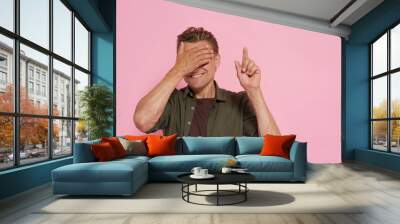 A man is wearing a green shirt and is pointing at something. He is smiling and has his hands covering his eyes Wall mural