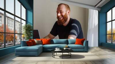 A man with a beard is sitting at a table with a laptop. He is smiling and laughing while using the laptop Wall mural