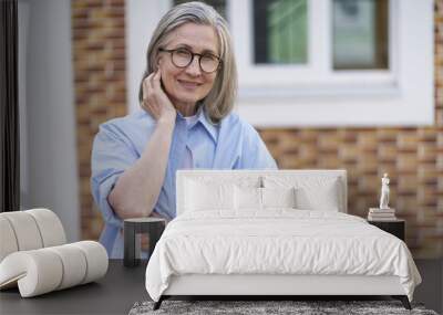 A woman in a blue shirt and glasses is smiling and leaning on a brick wall. She is happy and content Wall mural