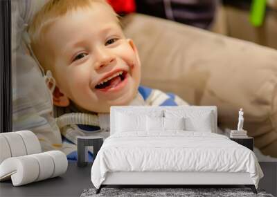 A young boy with a tracheostomy tube smiles brightly while sitting on a couch, radiating pure joy and infectious happiness. Wall mural