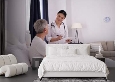 Beautiful nurse doing quick check up on patient. Cheerful friendly asian doctor holding clipboard to check on how her aged patient is doing. Wall mural