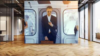 Businessman standing in subway and holding his bag. Young handsome man wearing full suit and round sunglasses standing in metro train and looking down at his phone he is holding in his hand. Wall mural