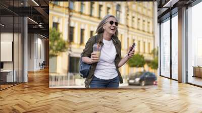 European senior woman with gray hair happy enjoying free time after work or traveling holding phone while having juice on the go using plastic cup in city background. Enjoying life mature woman Wall mural