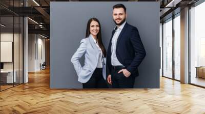 Two business partners stand confidently against a sleek gray background, sharing smiles as they engage with the camera, highlighting their professional camaraderie. Wall mural