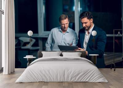 Two office collegues looking at tablet. Handsome businessman wearing suit showing something to his collegue on tablet late in evening. Wall mural