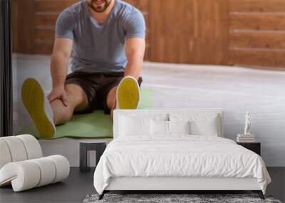 Young happy smiling fitness man doing stretching exercises at home. Amateur sports man in workout activity sitting on pad and stretch leaning to his toes. Hardworking guy exercising in apartment.  Wall mural