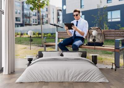 business, technology and people concept - young smiling businessman with tablet computer, headphones, backpack and electric scooter sitting on street bench in city Wall mural