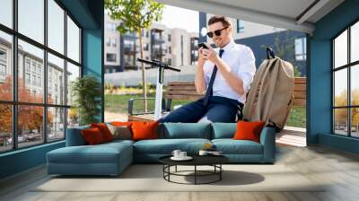 business and people and concept - young smiling businessman with smartphone, backpack and scooter sitting on street bench in city Wall mural