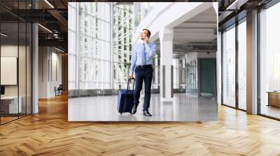 business trip, corporate and people concept - young businessman walking with travel bag along office building or airport and calling on smartphone Wall mural