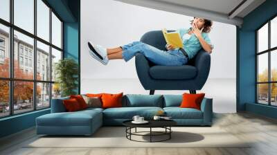 comfort, people and furniture concept - portrait of bored or tired young woman in turquoise shirt and jeans sitting in modern armchair and reading book and yawning over grey background Wall mural