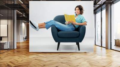 comfort, people and furniture concept - portrait of happy smiling young woman in turquoise shirt and jeans sitting in modern armchair and reading book over grey background Wall mural