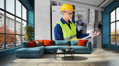 construction business and building concept - male electrician or worker in helmet and safety west with papers on clipboard and pencil at electric board Wall mural