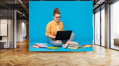 education, high school, technology and people concept - red haired teenage student girl in checkered shirt and torn jeans with books using laptop computer over bright blue background Wall mural