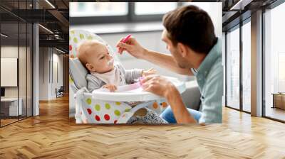 family, food, eating and people concept - father feeding little baby daughter sitting in highchair with puree by spoon at home Wall mural