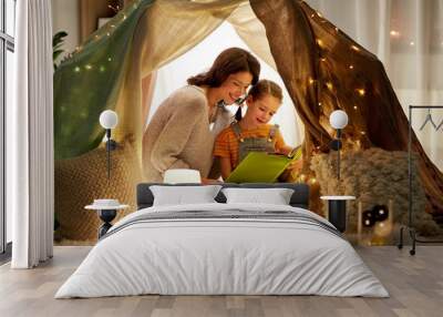 family, hygge and people concept - happy mother and little daughter reading book in kids tent at night at home Wall mural