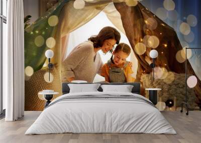 family, hygge and people concept - happy mother and little daughter reading book in kids tent at night at home Wall mural