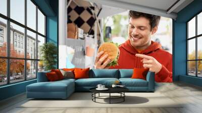 fast food, takeaway, eating and people concept - happy smiling young man in red hoodie showing hamburger over food truck on street background Wall mural