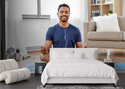 fitness, meditation and healthy lifestyle concept - indian man in earphones listening to music on smartphone and meditating in lotus pose at home Wall mural