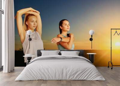 fitness, sport and healthy lifestyle concept - smiling young women or female friends with activity trackers stretching over sunset sky on background Wall mural