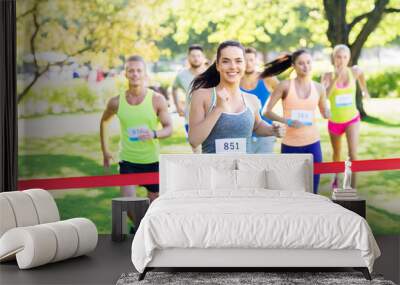 fitness, sport and success concept - happy teenage girl winning race and coming first to finish red ribbon over group of sportsmen running marathon with badge numbers at park Wall mural