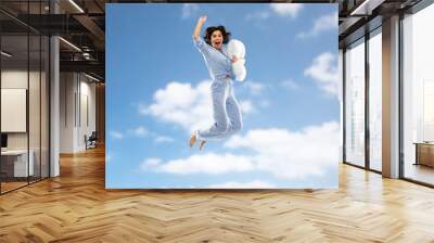 fun, people and bedtime concept - happy young woman full of energy in blue pajama holding pillow and jumping over blue sky and clouds background Wall mural