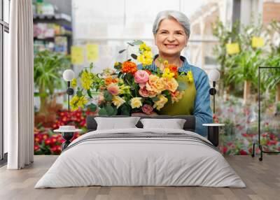 gardening, floristic and old people concept - portrait of smiling senior woman in green apron with bunch of flower over greenhouse at garden store on background Wall mural