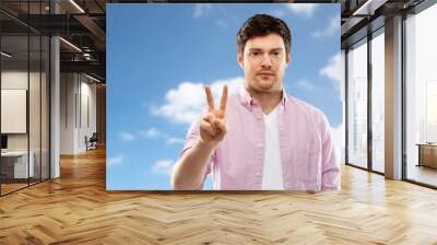 gesture, count and people concept - young man showing two fingers or peace hand sign over blue sky and clouds background Wall mural