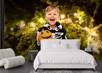 halloween, holiday and childhood concept - happy laughing boy in black costume with skeleton bones holding jack-o-lantern pumpkin over lights at night park on background Wall mural