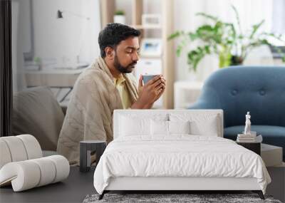 health, cold and people concept - sick young indian man in blanket drinking hot tea with lemon, honey and ginger at home Wall mural