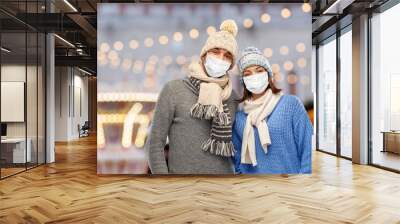 health, people and winter holidays concept - couple wearing face protective medical mask for protection from virus disease in knitted hats and scarves over christmas tree lights background Wall mural