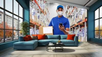 health, safety and pandemic concept - happy indian delivery man wearing face medical mask for protection from virus disease with smartphone and parcel box in uniform over warehouse background Wall mural