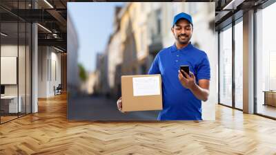 mail service, technology and shipment concept - happy indian delivery man with smartphone and parcel box in blue uniform over old tallinn city street background Wall mural