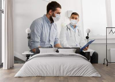 medicine, healthcare and pandemic concept - male doctor wearing face protective medical mask for protection from virus disease with clipboard and young man patient meeting at hospital Wall mural