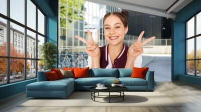 people, gesture and education concept - happy smiling teenage girl showing peace signs over city street or school yard background Wall mural