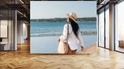 people, summer holidays and leisure concept - happy young woman in white shirt and straw hat with bag walking along beach Wall mural