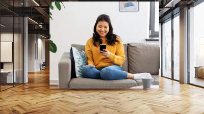 people, technology and leisure concept - happy smiling asian young woman in yellow sweater with smartphone sitting on sofa at home Wall mural