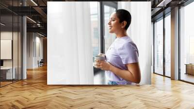 people and leisure concept - happy young african american woman with coffee cup looking out of window at home Wall mural