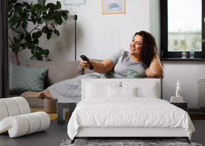 people and leisure concept - happy young woman lying on sofa with remote control and watching tv at home Wall mural