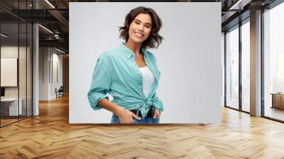 people concept - portrait of happy smiling young woman in turquoise shirt with hands in pockets over grey background Wall mural